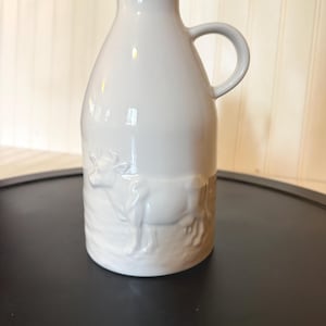 May include: A white ceramic milk jug featuring a raised, embossed cow design on the lower portion. The vessel has a narrow neck, a flared rim, and a single curved handle on the side. The surface is smooth and glossy, with a consistent white finish.