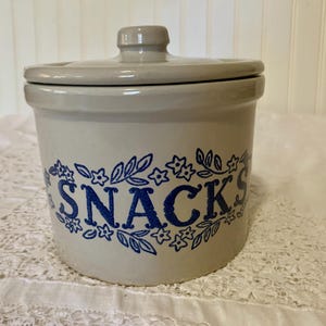 May include: A stoneware snack jar with a lid. The jar is light gray with a blue floral design around the word "SNACKS". The lid has a small knob for easy removal. A charming addition to any kitchen.