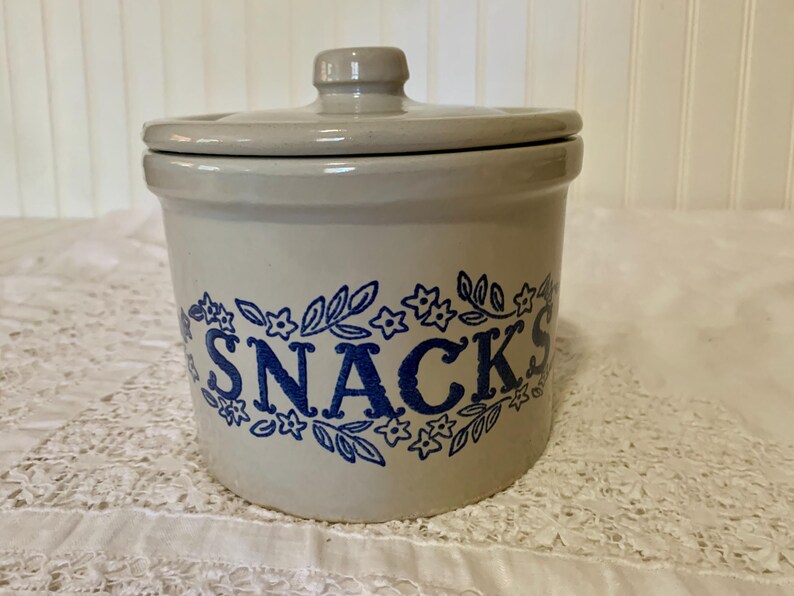 May include: A stoneware snack jar with a lid. The jar is light gray with a blue floral design around the word "SNACKS". The lid has a small knob for easy removal. A charming addition to any kitchen.