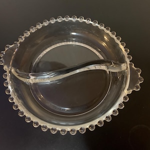 May include: Clear glass serving dish with a divided interior and a curved handle. The rim is decorated with small, round glass beads. The dish is designed for serving snacks or appetizers. The dish is on a dark surface.