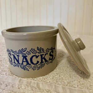 May include: A light beige ceramic snack jar with a cylindrical body and a matching lid. The front features the word SNACKS printed in bold blue letters, surrounded by a decorative blue floral and leaf pattern encircling the container.