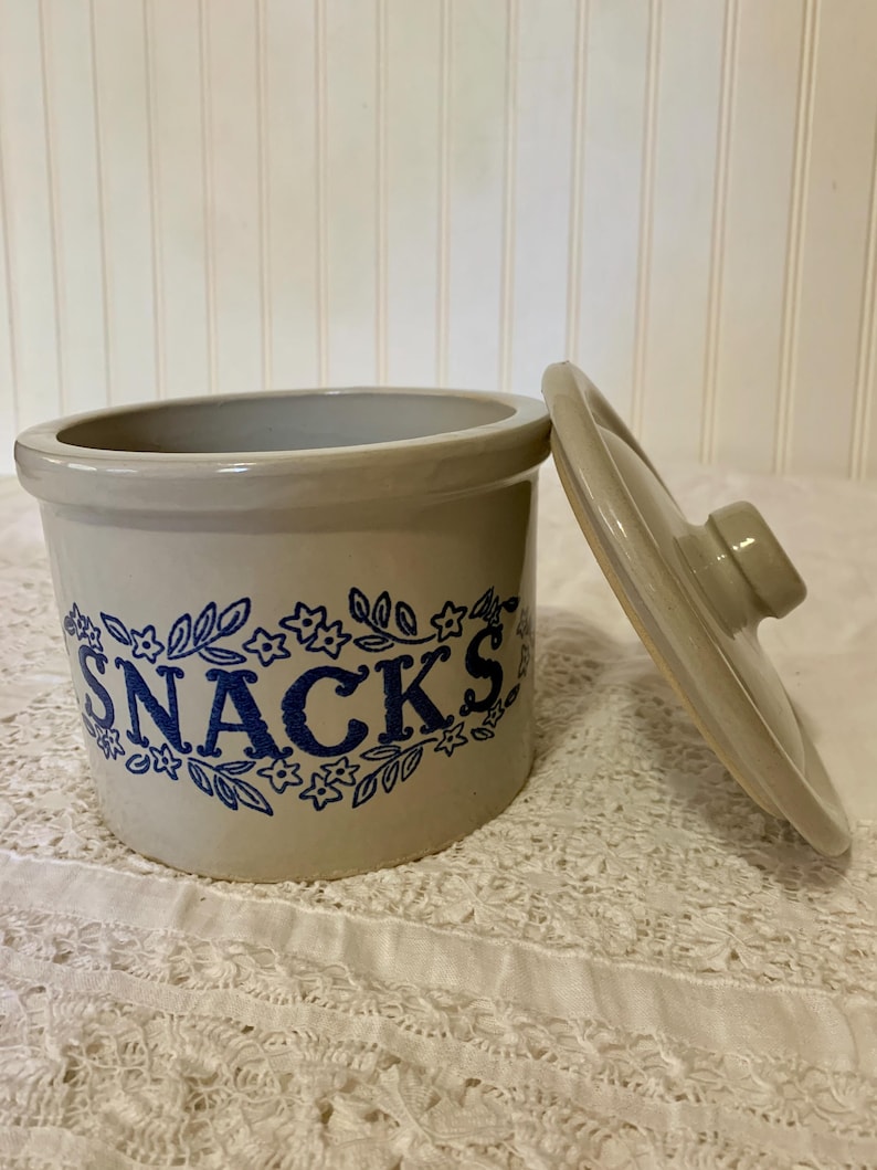 May include: A light beige ceramic snack jar with a cylindrical body and a matching lid. The front features the word SNACKS printed in bold blue letters, surrounded by a decorative blue floral and leaf pattern encircling the container.