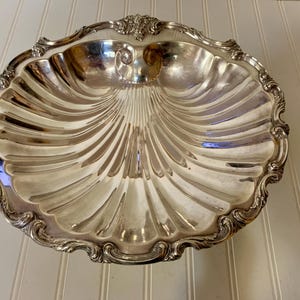 May include: A silver-colored, shell-shaped serving dish with a scalloped edge and ornate detailing. The dish has a ribbed interior and a decorative rim, reflecting light. This vintage piece is suitable for serving appetizers or displaying decorative items.