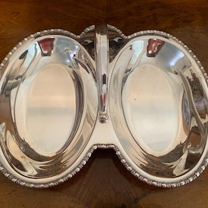 May include: A silver serving dish with two oval compartments and a handle. The dish has a beaded edge.