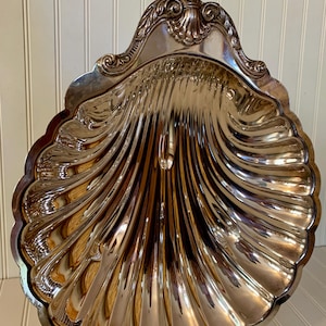 May include: A silver-colored, shell-shaped serving dish with a decorative, ornate rim. The dish has a scalloped edge and a ridged interior, reflecting light. The dish is supported by small, decorative feet.