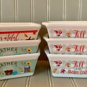 May include: Set of three white ceramic baking dishes with autumn-themed designs. The top dish features the word "grateful" in red script. The middle dish features the word "gather" in green script. The bottom dish features the word "gather" in green script. The second set of three dishes features the word "fall" in red script. The top dish has the word "fall" in red script. The middle dish has the word "fall is my" in red script. The bottom dish has the word "fall is my favorite color" in red script.
