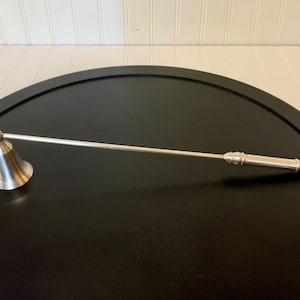 May include: A silver candle snuffer with a bell-shaped head and a long, slender handle. The snuffer is made of a shiny, metallic material and is designed to extinguish candles safely. The handle has decorative detailing near the end.