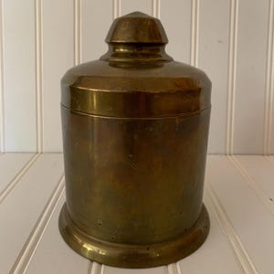 May include: A vintage brass jar with a lid. The jar has a rounded shape and a smooth, polished surface. The lid has a small knob on top.