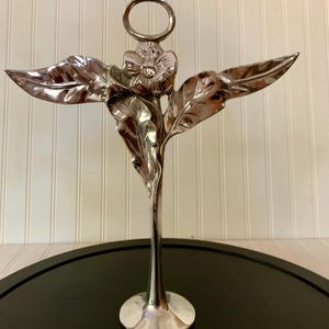 May include: A silver-toned metal stand featuring a central stem topped with a single flower and two extending leaves. A circular ring is positioned directly above the flower. The entire structure is supported by a circular base.
