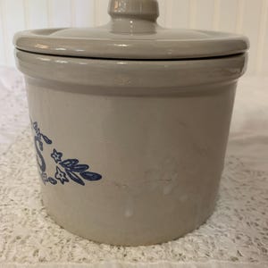 May include: A beige stoneware crock with a matching lid. The crock features a blue floral design on the side. The lid has a small knob for easy removal. The crock is likely used for food storage.