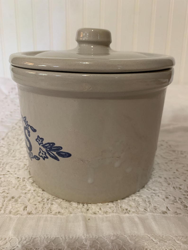 May include: A beige stoneware crock with a matching lid. The crock features a blue floral design on the side. The lid has a small knob for easy removal. The crock is likely used for food storage.