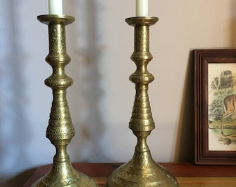Könnte beinhalten: Zwei verzierte goldene Kerzenleuchter mit weißen Kerzen. Die Kerzenleuchter haben ein detailliertes Design und eine breite Basis.