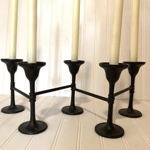 May include: A black metal candelabra with five candle holders, each holding a cream-colored candle. The candelabra has a unique design with connecting bars between the holders, creating a decorative centerpiece.