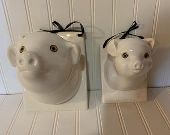 May include: Two white ceramic pig wall hangings. One is a pig head, the other a piglet head. Both have black ribbon for hanging and golden-brown eyes. The pig heads are mounted on rectangular plaques.