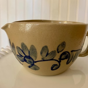 May include: Tan stoneware mixing bowl with a handle and spout. The bowl features a blue floral design around the exterior. The bowl is sitting on a white, scalloped-edge plate.