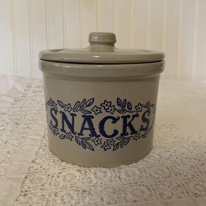 May include: A beige ceramic snack jar with a lid. The word "SNACKS" is printed in blue script, surrounded by a floral design. The jar is cylindrical and sits on a white lace tablecloth.