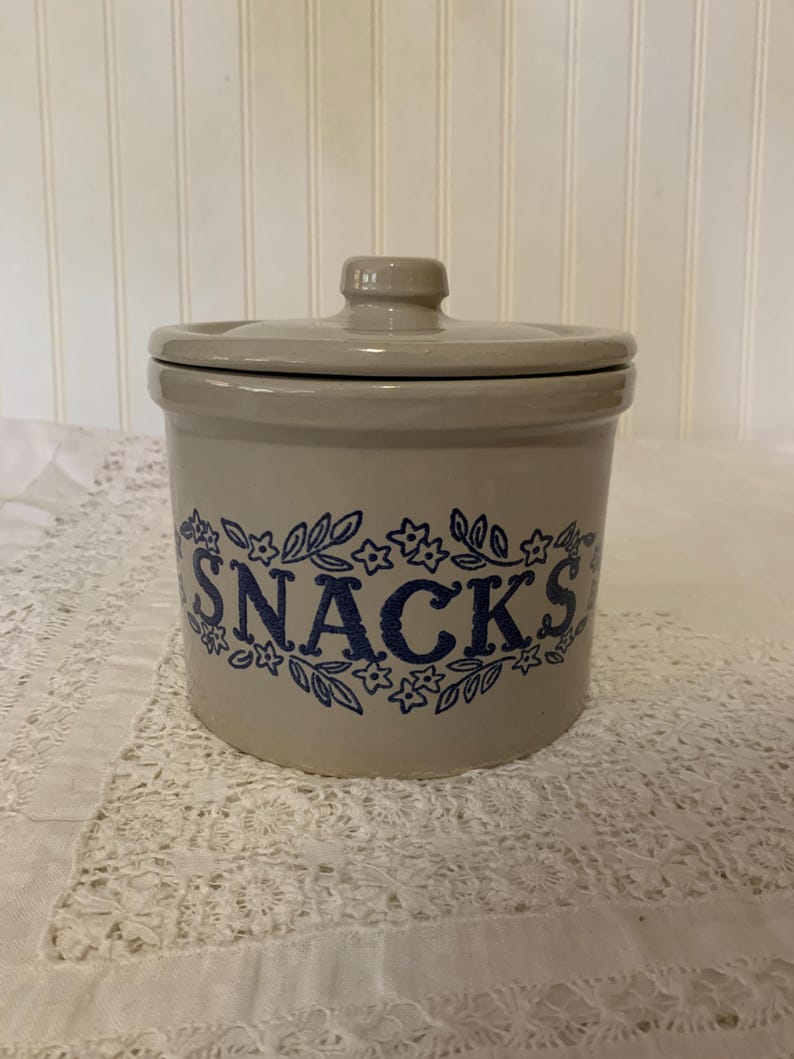 May include: A beige ceramic snack jar with a lid. The word "SNACKS" is printed in blue script, surrounded by a floral design. The jar is cylindrical and sits on a white lace tablecloth.