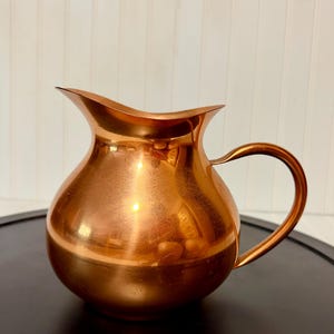 May include: A polished copper pitcher with a rounded body, a wide pouring spout, and a curved handle. The metallic surface reflects light, showcasing a warm, reddish-brown tone and a smooth, reflective finish across the entire exterior of the vessel.