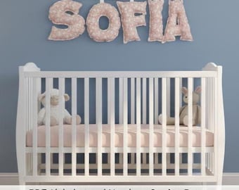 May include: A white crib with a pink mattress and two stuffed animals. Above the crib, the name "SOFIA" is spelled out in large, floral-patterned letters. The letters are a light pink color and are suspended from a white string against a blue wall.