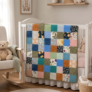 May include: A patchwork quilt featuring a grid of square fabric panels in various colors and patterns, including shades of blue, green, orange, pink, and floral prints. The quilt is draped over the side of a white wooden crib in a nursery setting.