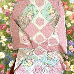 Può includere: Una trapunta patchwork con quadrati alternati di tessuto a stampa floreale nei toni del rosa, blu e bianco, accentuata da applicazioni di fiori in tessuto tridimensionali. Il testo in basso recita: Shabby Flower Baby Quilt Pattern and Instructions. PDF Digital Download.