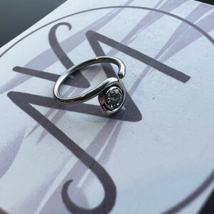 Puede incluir: Un anillo de plata con una piedra de cristal transparente. El anillo est&aacute; sobre un fondo blanco con un dise&ntilde;o de espiral gris.