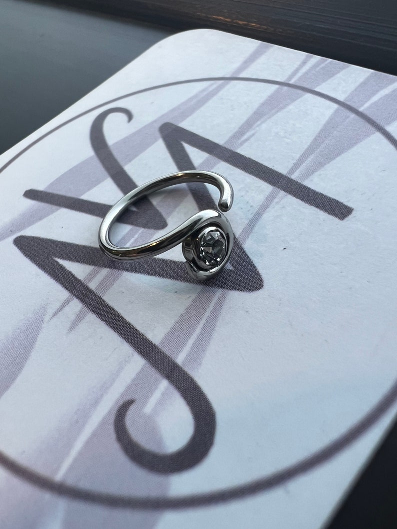 Puede incluir: Un anillo de plata con una piedra de cristal transparente. El anillo est&aacute; sobre un fondo blanco con un dise&ntilde;o de espiral gris.