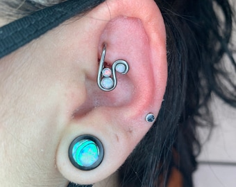 Puede incluir: Primer plano de la oreja de una persona con un piercing de hélice plateado con piedras de ópalo. El piercing tiene la forma de una línea curva con dos piedras de ópalo. La persona también tiene un dilatador de oreja de concha abulón negra y verde en su oreja inferior.