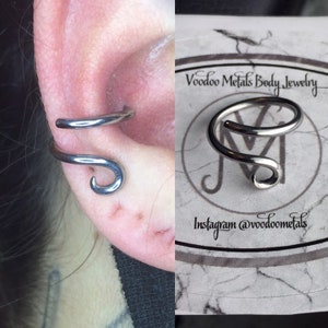 May include: A silver ear cuff with a curved design. The cuff is shown on a person's ear and also on a white card with the text "Voodoo Metals Body Jewelry" and "Instagram @voodoometals".