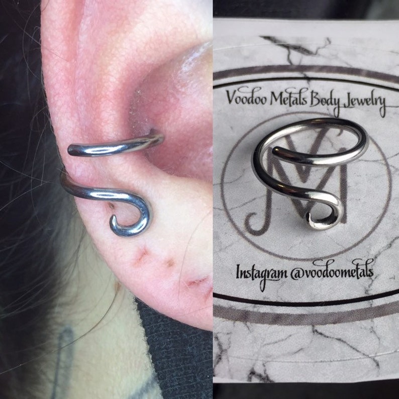 May include: A silver ear cuff with a curved design. The cuff is shown on a person's ear and also on a white card with the text "Voodoo Metals Body Jewelry" and "Instagram @voodoometals".