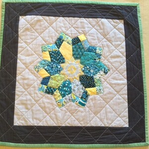 May include: A small, quilted throw pillow with a green border and a black inner border. The center of the pillow features a colorful patchwork design in shades of blue, green, and yellow.