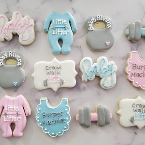 May include: A set of 12 decorated sugar cookies with a baby theme. The cookies feature various designs including a baby onesie, a dumbbell, a kettlebell, and the words "Baby", "Little Lifter", "Crawl Walk", and "Burpee Machine".