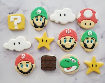 Könnte beinhalten: Ein Set mit 11 dekorierten Zuckerkeksen mit Figuren und Objekten aus dem Super Mario Bros. Videospiel-Franchise. Die Kekse sind mit grünem, rotem, gelbem und weißem Zuckerguss verziert. Die Figuren sind Mario, Luigi und Yoshi. Die Objekte sind ein Fragezeichenblock, ein Stern und eine Wolke.