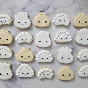 May include: A collection of 20 decorated sugar cookies shaped like dumplings. The cookies are white, beige, and light brown with black eyes and smiles.