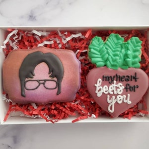 May include: Two decorated sugar cookies in a white box with red and white shredded paper. One cookie is a portrait of a man with black hair and glasses. The other cookie is a heart-shaped beet with green leaves and the text "my heart beets for you".