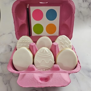 May include: A pink egg carton filled with six white sugar cookies decorated with icing. The cookies are shaped like Easter eggs and have different designs. A small card with four colored circles is visible in the carton.