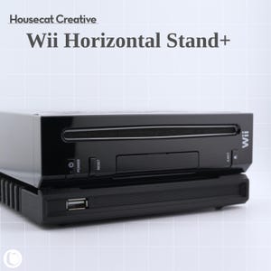 Puede incluir: Una consola Wii negra sobre un soporte horizontal negro. El soporte tiene un puerto USB y una superficie texturizada. La consola tiene botones de encendido, reinicio y expulsión. El texto "Wii Horizontal Stand+" es visible.
