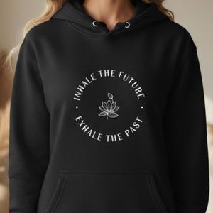 May include: Black hoodie with a front pocket and drawstring hood. The hoodie features a white circular graphic with the text "INHALE THE FUTURE, EXHALE THE PAST" and a lotus flower illustration. The hoodie is made of a soft material.