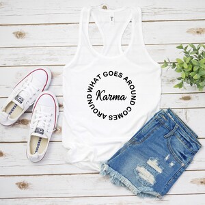May include: White tank top with the text "Karma" in a circular design. The image also includes a pair of white sneakers with red trim and denim shorts with distressed details. The items are arranged on a light wood-grain surface.