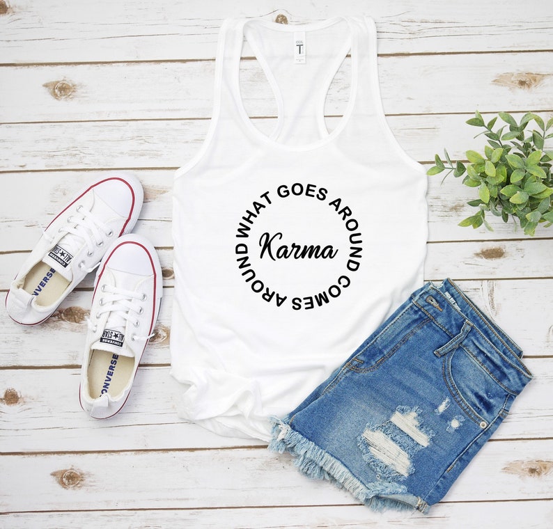 May include: White tank top with the text "Karma" in a circular design. The image also includes a pair of white sneakers with red trim and denim shorts with distressed details. The items are arranged on a light wood-grain surface.