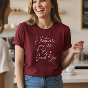 Op de afbeelding: Een bordeauxrood T-shirt met opgerolde mouwen. Het shirt heeft de witte tekst 'Whatever you are, Be a Good One'. De persoon draagt een blauwe spijkerbroek. De achtergrond is wazig.