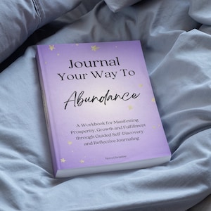May include: A hardback book titled "Journal Your Way To Abundance" with a light purple cover and gold star accents. The book's title is in a script font. The book is on a grey fabric surface.