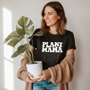 K&ouml;nnte beinhalten: Dunkelgraues T-Shirt mit dem wei&szlig;en Schriftzug "PLANT MAMA". Die Person tr&auml;gt eine beige Strickjacke und blaue Jeans. Eine Topfpflanze mit gro&szlig;en gr&uuml;nen Bl&auml;ttern wird in einem wei&szlig;en Topf gehalten.