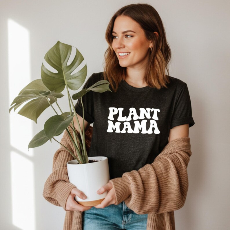 K&ouml;nnte beinhalten: Dunkelgraues T-Shirt mit dem wei&szlig;en Schriftzug "PLANT MAMA". Die Person tr&auml;gt eine beige Strickjacke und blaue Jeans. Eine Topfpflanze mit gro&szlig;en gr&uuml;nen Bl&auml;ttern wird in einem wei&szlig;en Topf gehalten.