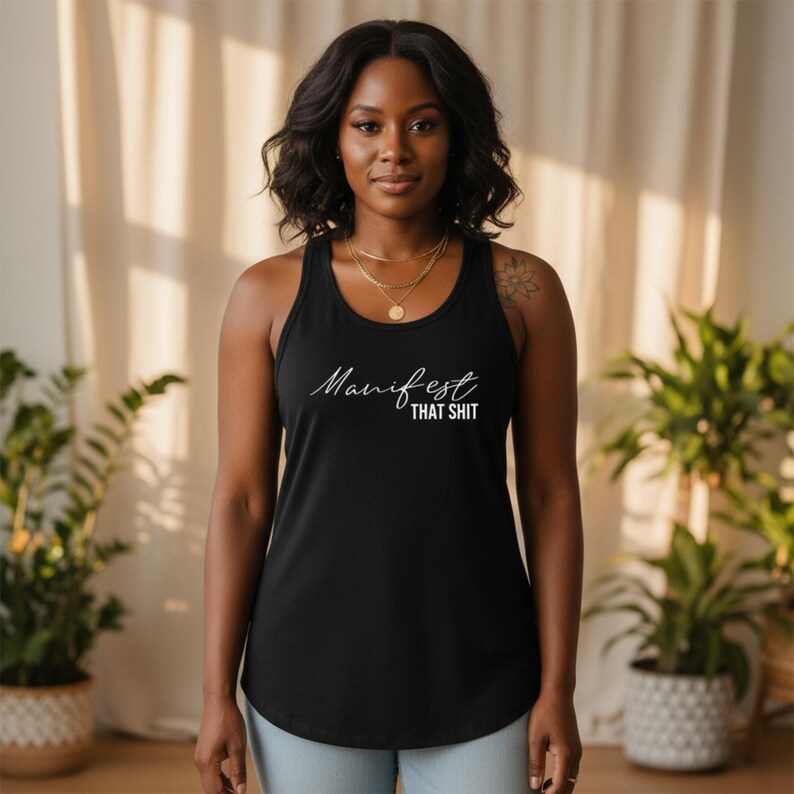 May include: Black racerback tank top with the words "Manifest That Shit" in white script. The tank top is made of a soft material and has a rounded hemline. The model is wearing gold necklaces.