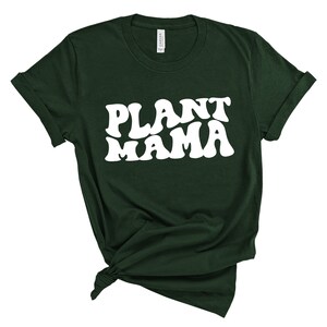 K&ouml;nnte beinhalten: Dunkelgr&uuml;nes T-Shirt mit dem wei&szlig;en Text "PLANT MAMA" in einem Retro-Schriftzug. Der Saum des T-Shirts ist geknotet und die &Auml;rmel sind hochgekrempelt. Das T-Shirt ist aus weichem Material.
