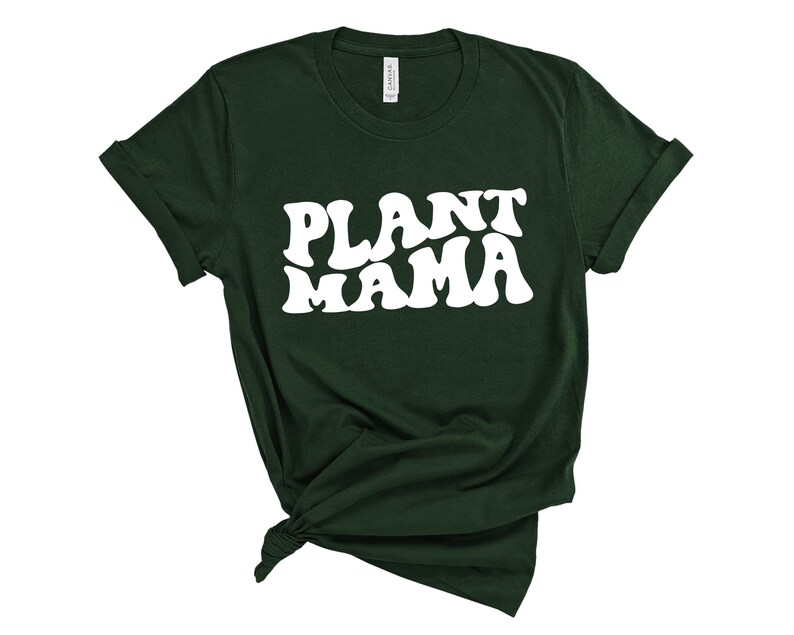 K&ouml;nnte beinhalten: Dunkelgr&uuml;nes T-Shirt mit dem wei&szlig;en Text "PLANT MAMA" in einem Retro-Schriftzug. Der Saum des T-Shirts ist geknotet und die &Auml;rmel sind hochgekrempelt. Das T-Shirt ist aus weichem Material.