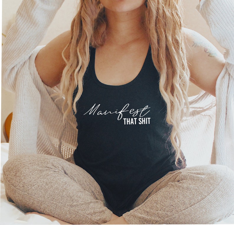 May include: Black tank top with the words "Manifest That Shit" in white script. The top is a scoop neck style. The person is wearing a white cardigan and tan pants.