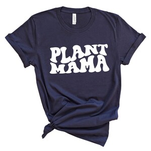 K&ouml;nnte beinhalten: Marineblaues T-Shirt mit dem wei&szlig;en Text "PLANT MAMA" in einem Retro-Schriftstil. Das Shirt ist am Saum geknotet und hat hochgekrempelte &Auml;rmel. Das T-Shirt besteht aus einem weichen Material.