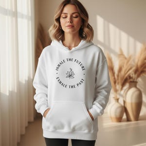 May include: White hoodie with a front pocket and drawstring. The hoodie features a circular graphic with the text "INHALE THE FUTURE, EXHALE THE PAST" surrounding a lotus flower design. The model is wearing black pants.
